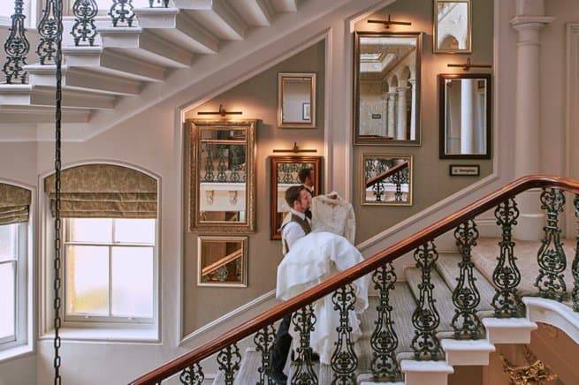 A man carrying a wedding dress up grand staircase at a British country house wedding venue.