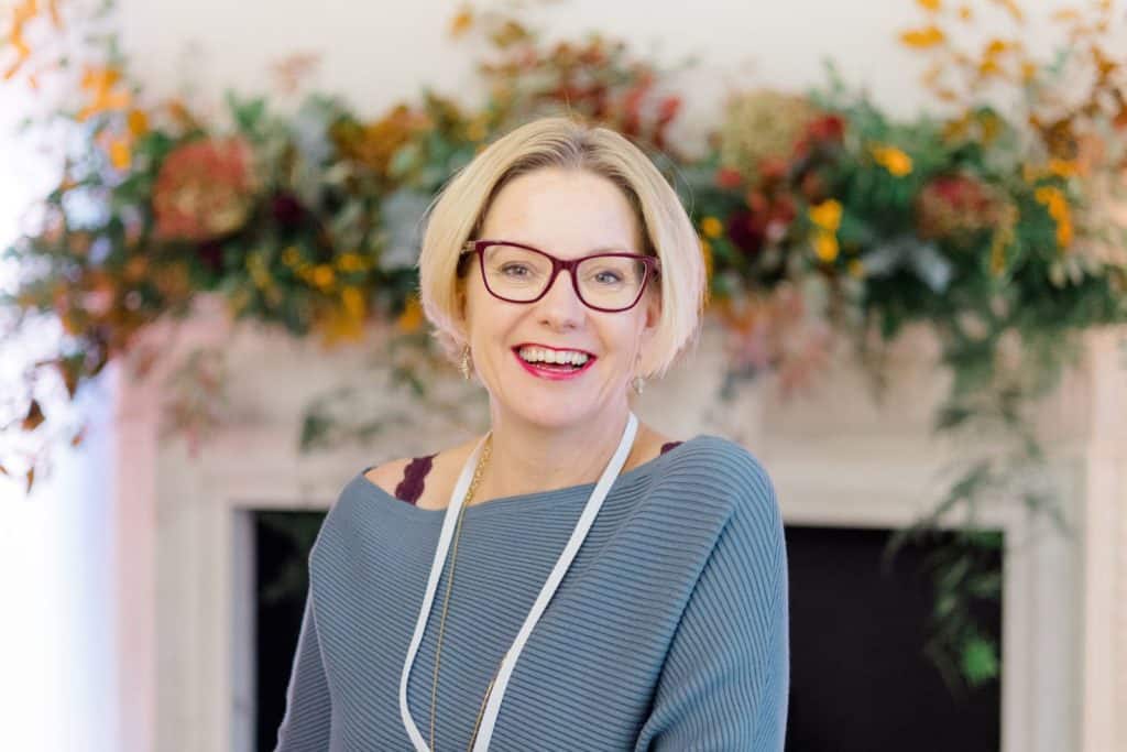Woman wearing blue jumper standing in front of flowers smiling at camera | Kelly Chandler Success Coach